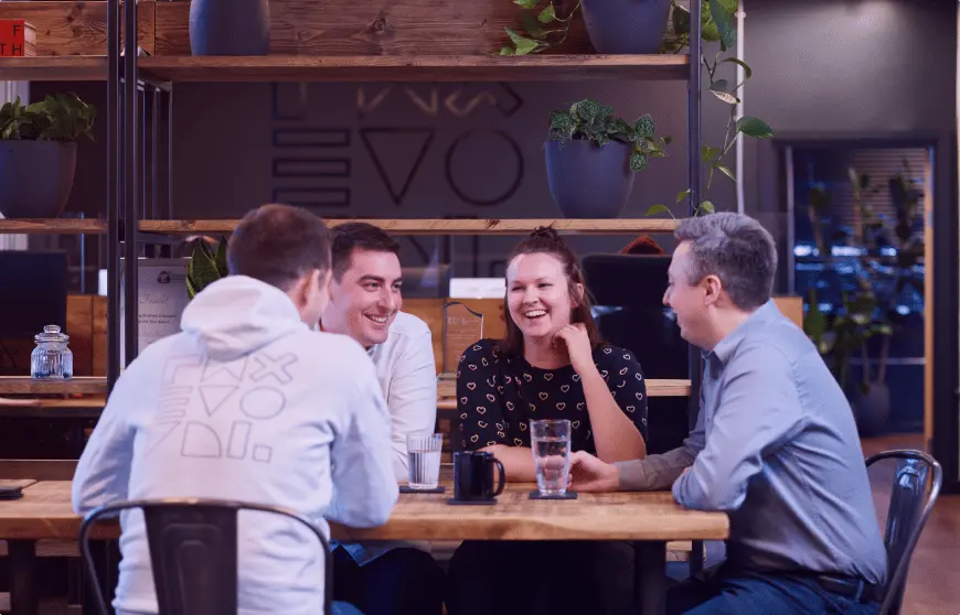 Four Evo Agency employees sitting at a table together in the centre of the office talking and smiling.